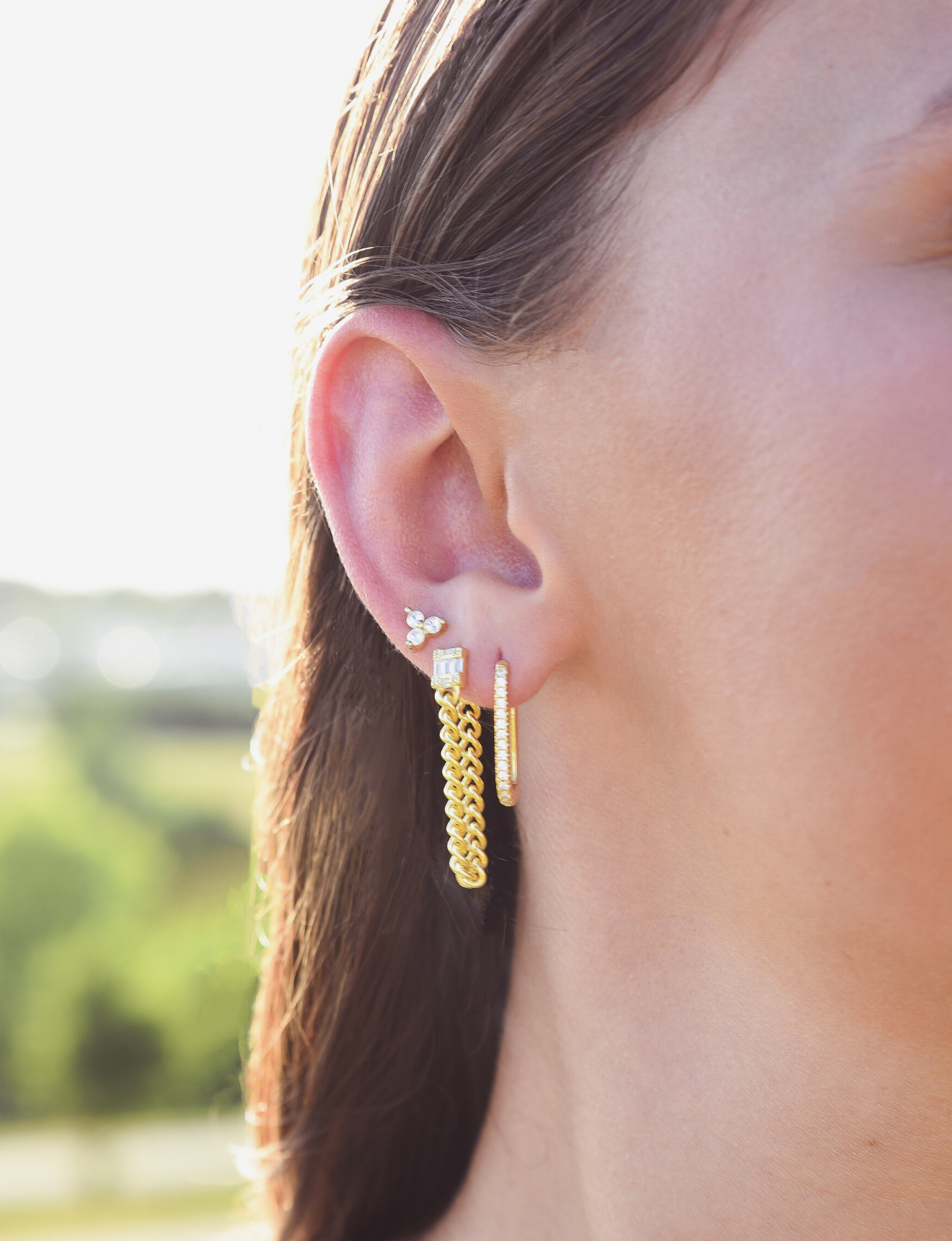 Side of Burdlife Model's head displaying 3 earrings including the Luxe Oval Hoops, the Refined Single Chain Earring and the Modish Triple Crystal Studs. 