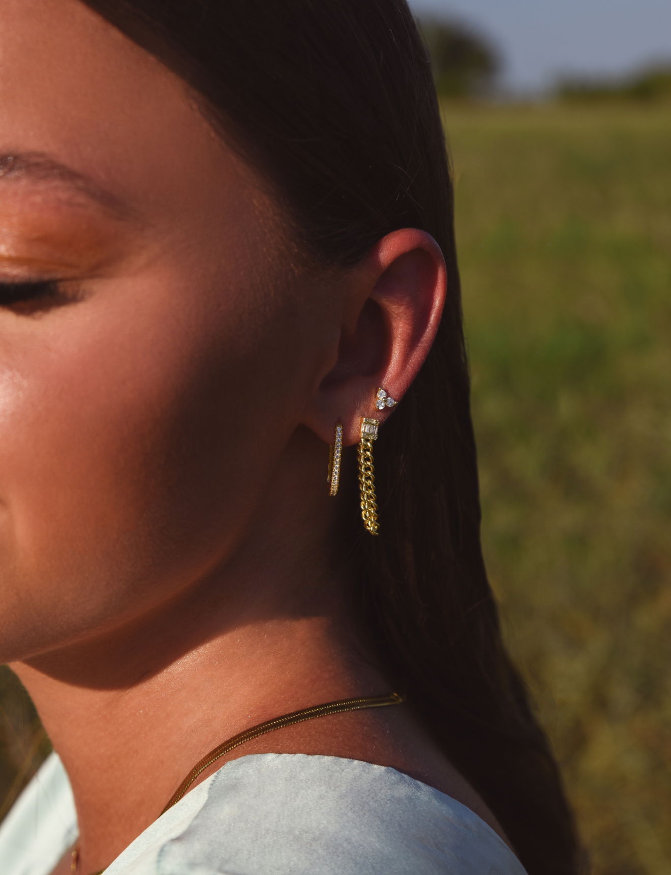 Side of Burdlife Model's head displaying 3 earrings including the Luxe Oval Hoops, the Refined Single Chain Earring and the Modish Triple Crystal Studs. 